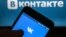  A hand holding a smarthphone showing the logo of the VK (Vkontakte) social network, in front of a computer screen