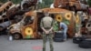 Военнослужащий смотрит, как художник Трек Келли раскрашивает автомобиль, сожженный во время вторжения России в Украину 10 августа 2022 года