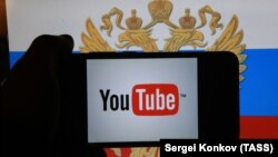A hand holding a smartphone with YouTube's logo on the screen, in front of a computer screen in St. Petersburg, Russia showing the Russian national flag and coat of arms