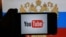 A hand holding a smartphone with YouTube's logo on the screen, in front of a computer screen in St. Petersburg, Russia showing the Russian national flag and coat of arms